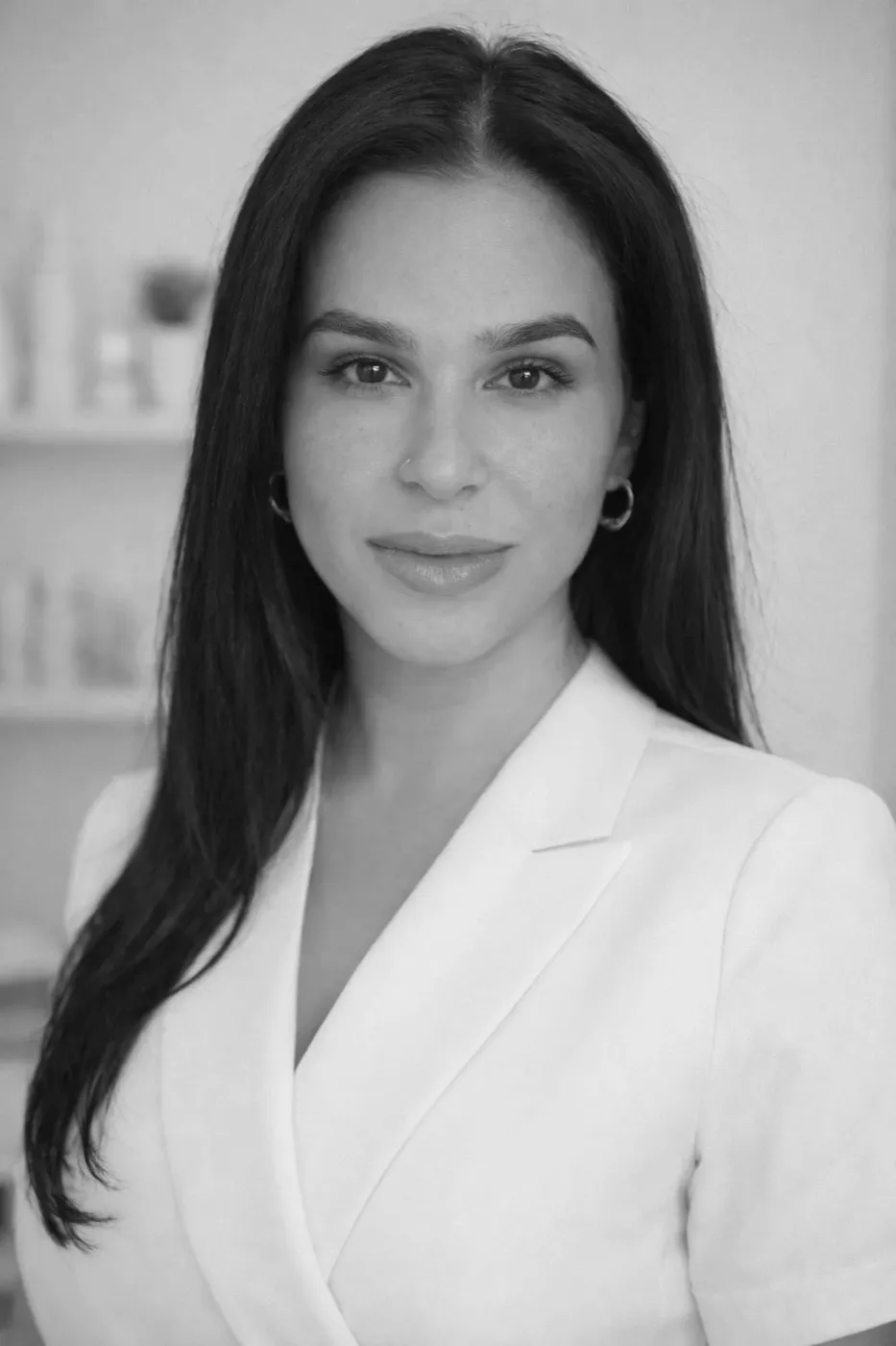 Alena, a licensed esthetician, in a white jacket in front of a shelf of skincare products.